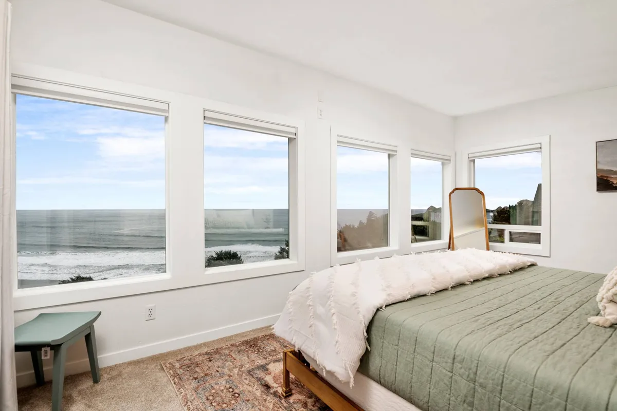 A peaceful bedroom featuring a large bed with soft bedding positioned near expansive windows that offer a stunning ocean view. The decor is serene and inviting, with a decorative mirror reflecting natural light, creating a relaxing retreat perfect for unwinding or reading.