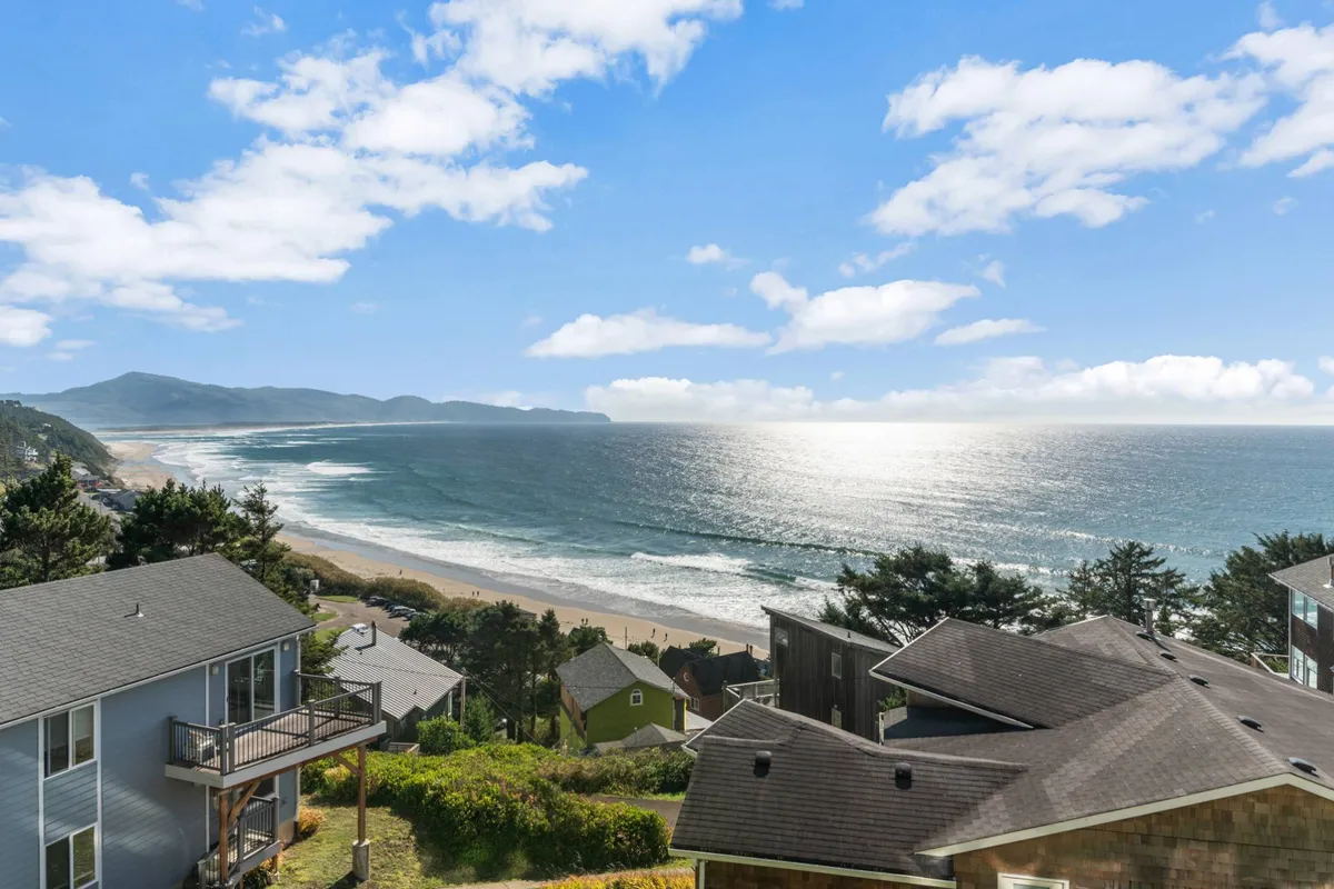 A breathtaking coastal view featuring rolling ocean waves, distant mountains, a cloudy sky, and coastal houses nestled along the shoreline. The scene exudes serenity and showcases the natural beauty visitors can expect from the Oregon Coast.