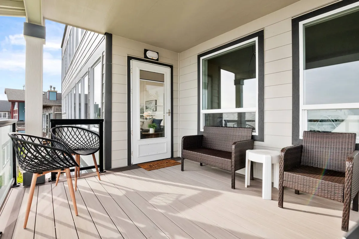 An inviting outdoor patio area with wicker chairs positioned near a white door and a decorative mat, suggesting a space for relaxation, reading, or socializing. The calm, welcoming mood hints at evenings spent under the stars or casual drinks outdoors.