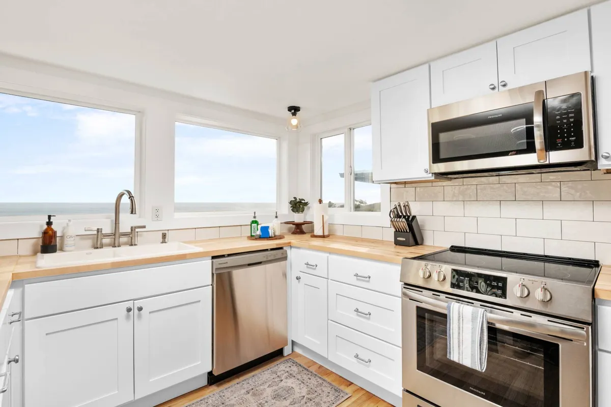 A modern kitchen with sleek appliances, warm wooden countertops, and large windows that frame a spectacular ocean view. The space is bathed in natural light, with a sink situated perfectly to enjoy the scenery while cooking or cleaning. The inviting atmosphere is accentuated by the cheerful decor and the seamless connection to the outdoors.
