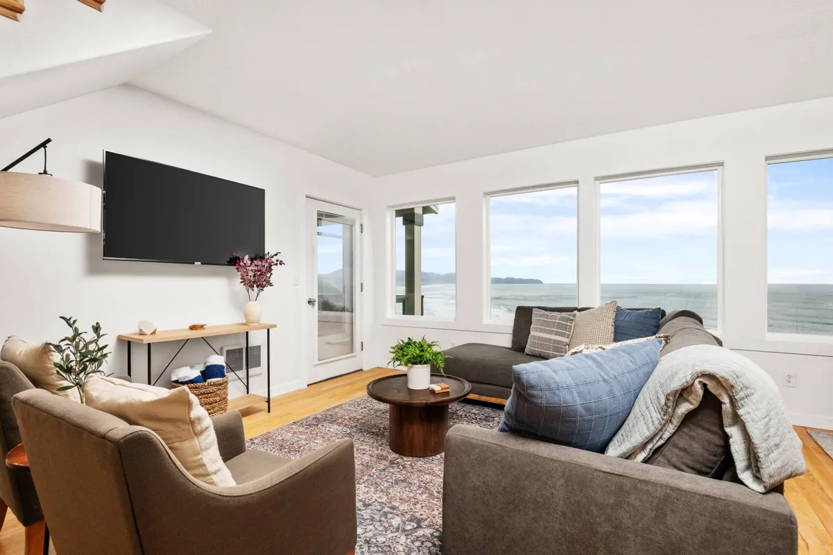 The image captures a cozy living room with a plush sofa facing expansive windows that frame stunning ocean views. Natural light fills the space, highlighting a stylish rug and inviting seating area. A wall-mounted TV is visible, but the primary focus is on the tranquil ambiance and the breathtaking seascape beyond the windows.