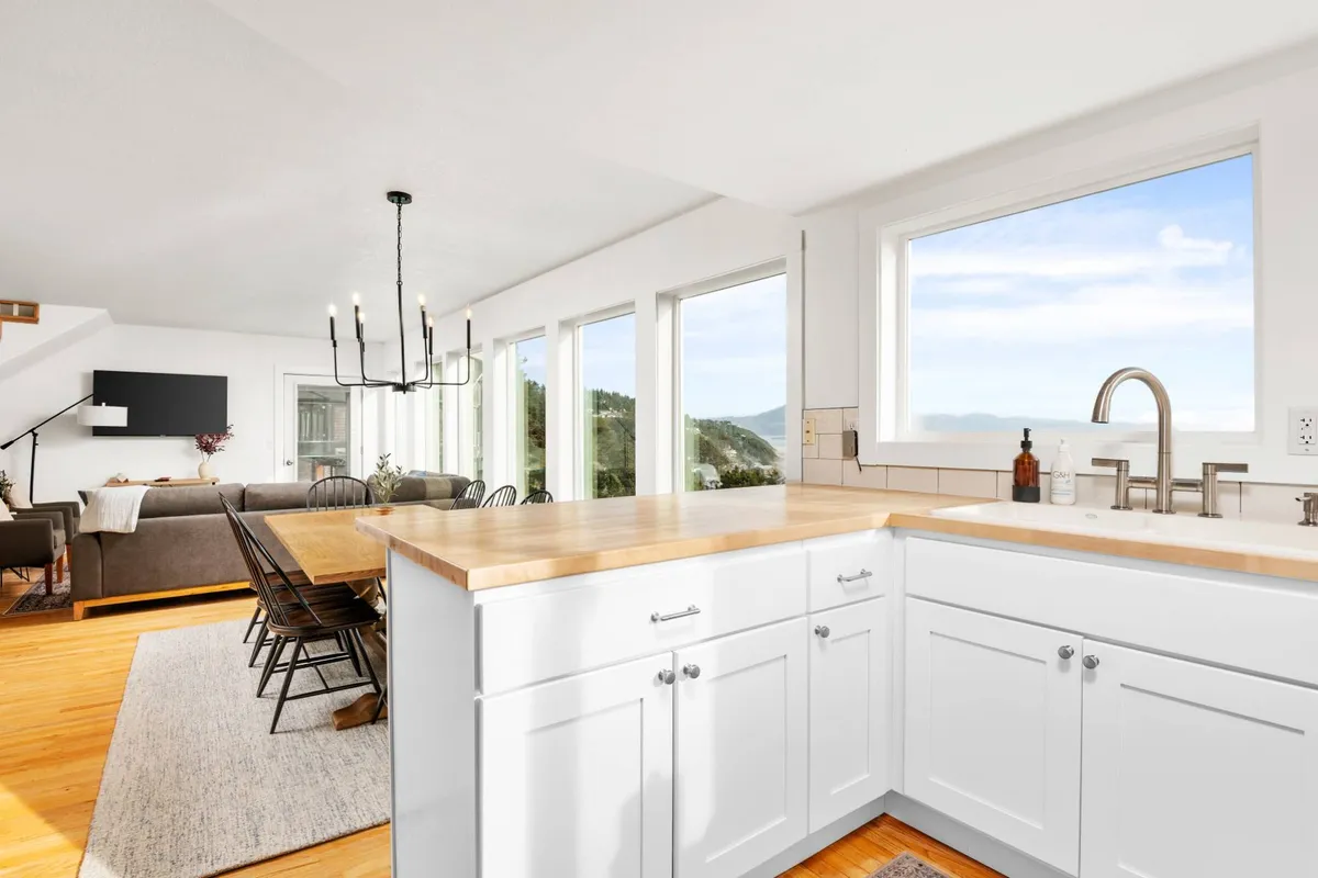 An inviting interior scene featuring a spacious kitchen island, stainless steel appliances, and a dining area set against large windows. The dining table is ideally positioned for group gatherings, with a chandelier overhead and expansive mountain or ocean views visible beyond the glass.