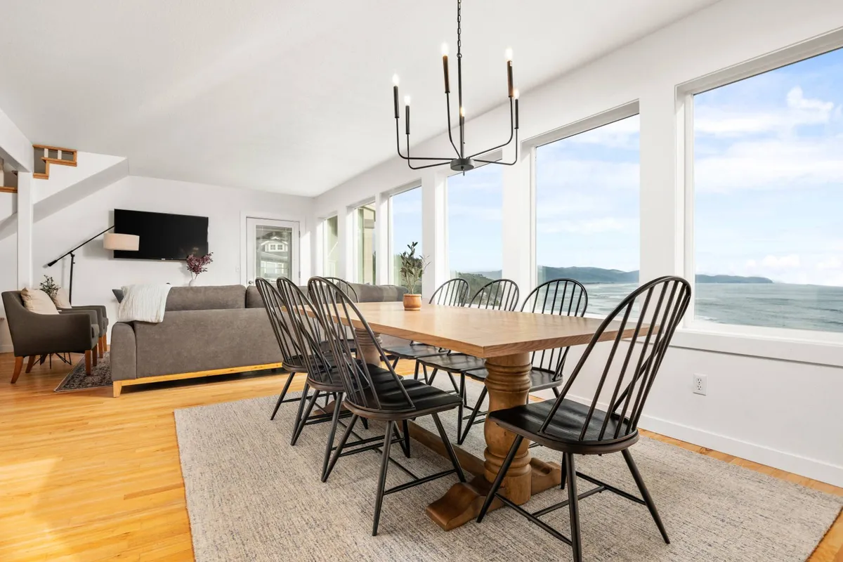 This interior image features a spacious and inviting dining area with a large table, comfortable seating, a striking chandelier overhead, and expansive windows that frame scenic coastal views. The furniture and décor are modern yet cozy, creating a perfect setting for gathering and socializing.