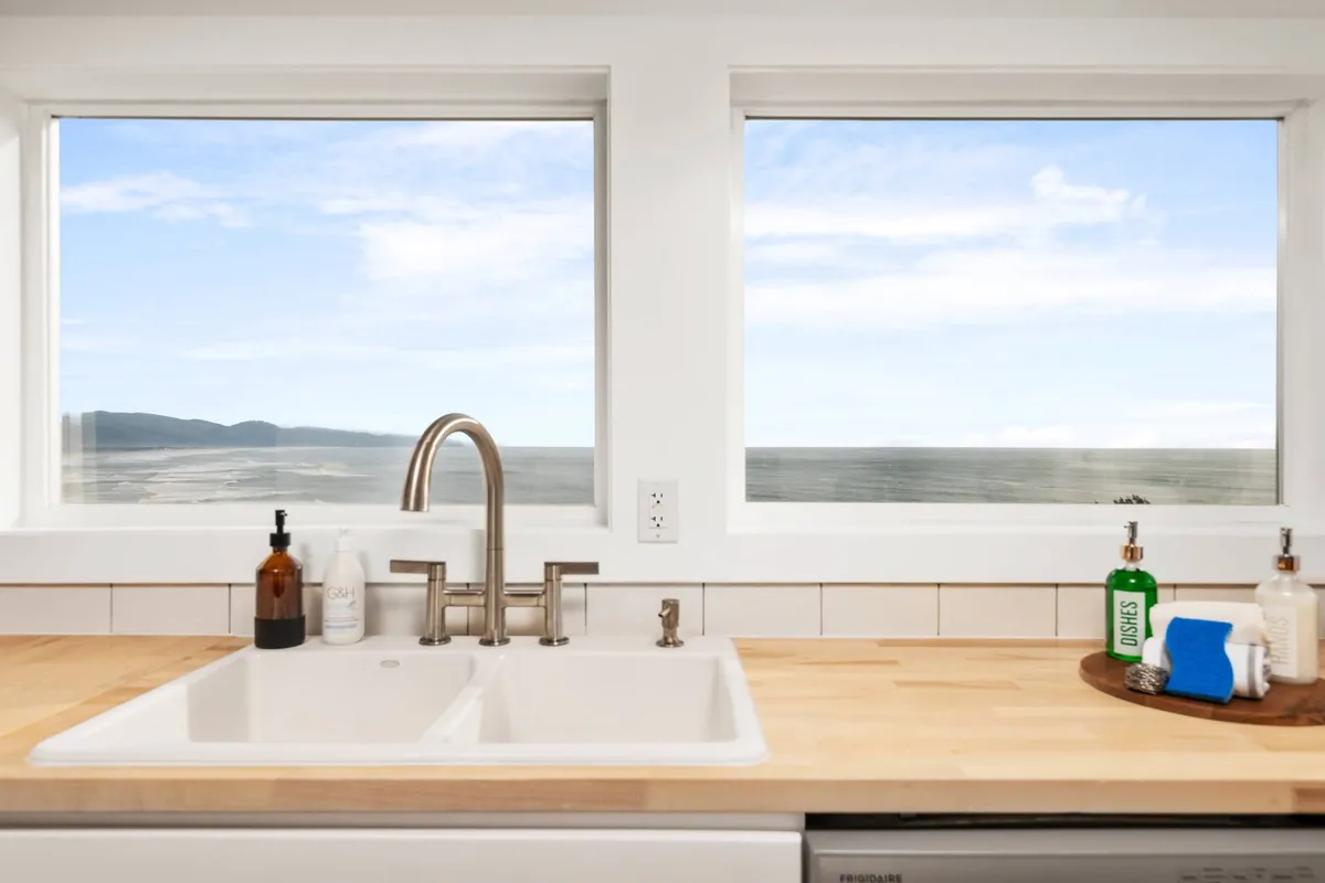 A sunlit kitchen scene where a sleek sink and wooden countertop sit beneath large windows, which offer stunning ocean views. The area is bright, modern, and designed for both cooking and socializing, with the beauty of the Pacific just outside.